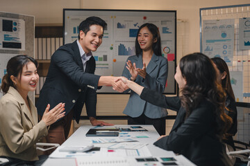 Two business people are shaking hands after closing a deal in a meeting room, while their colleagues are clapping and smiling, celebrating the achievement