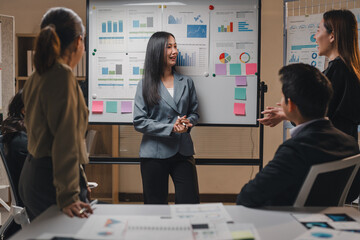 Asian businesswoman leading a meeting with her colleagues, presenting charts and graphs on a whiteboard using sticky notes, discussing business strategies and performance