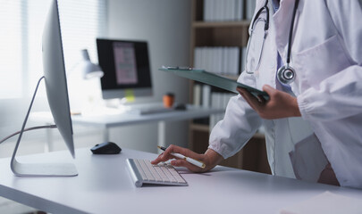 Female physician using computer keyboard and holding patient medical records in bright modern clinic or hospital office, typing medical report