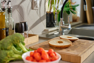 Sliced bagel on wooden cutting board with cherry tomatoes, broccoli and kitchen tools in background. Preparation of healthy breakfast or snack in home kitchen