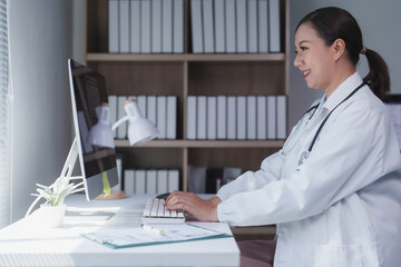 Asian female doctor wearing lab coat and stethoscope is working on desktop computer in her bright...