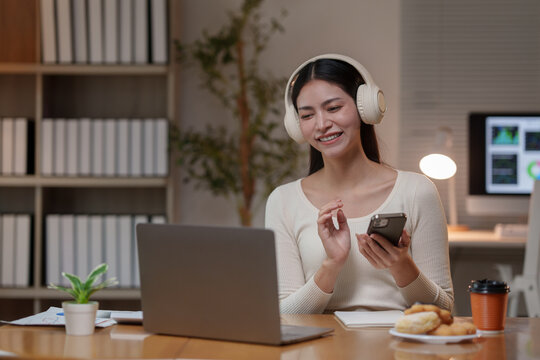 Young beautiful asian woman wearing headphones working with laptop and using smartphone while sitting at her desk in home office at night, working or studying online, e-learning concept
