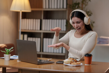 Young asian businesswoman wearing headphones is showing the size of something with her hands while making a video conference on a laptop, sitting at the desk in home office with pastries and coffee
