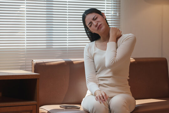 Young woman massaging her neck with a pained expression, experiencing discomfort from stiff muscles after prolonged hours of sedentary work at home on the sofa