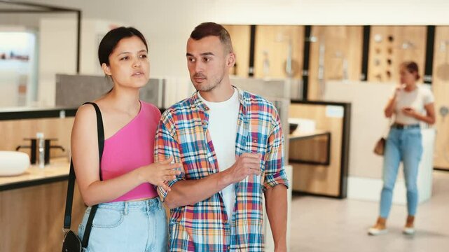 Interested young couple, man and woman of different ethnicities, amicably discussing choice of bathroom fixtures and furnishing during visit to specialized plumbing store