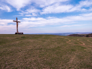 A wooden crucifix on a wide hilltop with several winding paths in the grass leading to it. A...