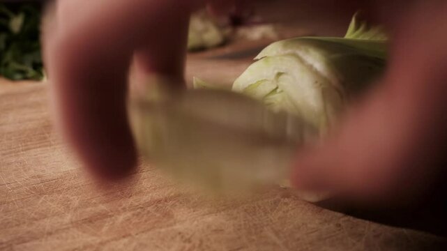 Side view of hands slicing fresh Pak Choi on a wooden board, cutting the end of the vegetable. Close-up food prep in warm kitchen light with leafy greens blurred in the background.