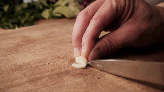 Close-up side view of garlic being finely chopped on a wooden board. Warm kitchen lighting, hand and knife in focus, leafy greens softly blurred in background.