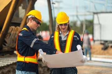 Two construction workers in hard hats and reflective vests discussing blueprints at an industrial...