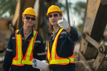 Two construction workers in hard hats and reflective vests discussing blueprints at an industrial...