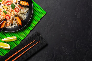 Fried rice with seafood mussels, shrimps and basil in a black plate with chopsticks on green bamboo mat and stone table