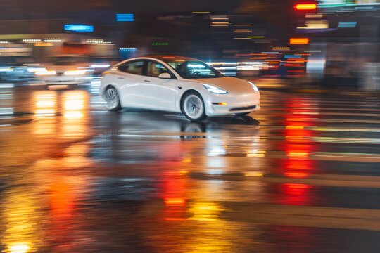 LOS ANGELES, CA - March 6, 2025: Tesla electric car rushing through city traffic, crossing illuminated wet street intersection on a rainy night in West Hollywood, California.