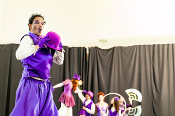 Comedian wearing purple clothes and bowler hat performing a funny sketch on stage with other actors in the background