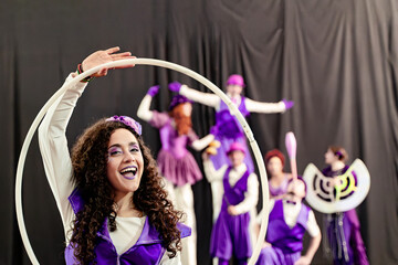 Young woman performing with hula hoop, smiling, during a circus show with other blurred artists in the background