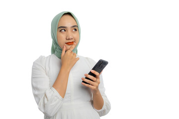 Fototapeta premium Pensive young Asian woman in green hijab and white blouse holding mobile phone, touching her chin, looking at copy space, thinking about online offers isolated on transparent background