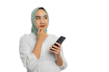 Pensive young Asian woman in green hijab and white blouse holding mobile phone, touching her chin, looking at copy space, thinking about online offers isolated on transparent background