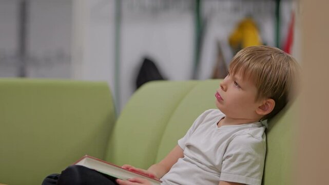 Static view of young blonde boy bored on the sofa holding a school book