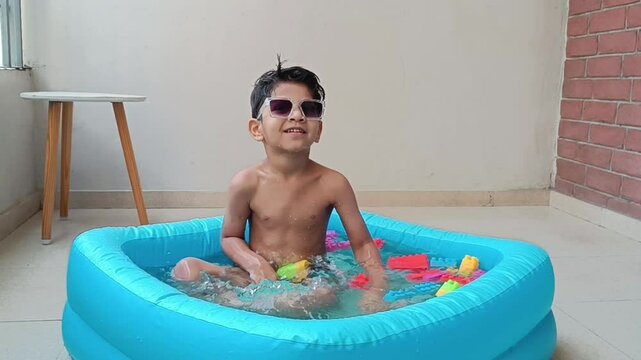 4 year old cute, young Indian boy relaxing in a kiddie pool- bath tub and splashing water, wearing stylish sunglasses and enjoying a fun indoor summer activity during summer vacation.