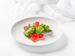 A gourmet dish of white fish fillet with roasted broccoli, cherry tomatoes, and red bell pepper, garnished with fresh basil. Served on a minimalist white plate against a clean, bright white background