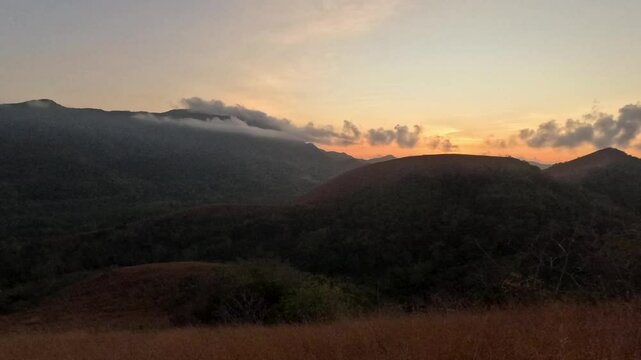 Sunrise glow appears behind mountain ridges near Mount Tapyas with misty clouds dry grass and dark hillside silhouettes in Coron Palawan