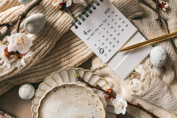 Fototapeta premium April 2025 calendar with April 20 marked, next to a golden pen, cup of coffee, blooming spring branches, and decorative Easter eggs on a knitted beige fabric background