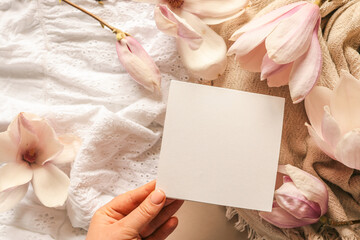 Blank square card on a soft neutral background surrounded by blooming magnolia flowers and lace...