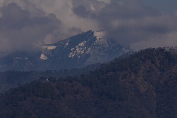A moody Himalayan landscape featuring snow covered mountain peaks above dense forested hills during the spring season in Himachal Pradesh, India.
