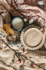 Flat lay with a cup of coffee and an unlit soy candle in a dark jar. Surrounded by cherry blossoms, pastel eggs, soft knit fabric, and a floral dress — cozy spring morning vibe