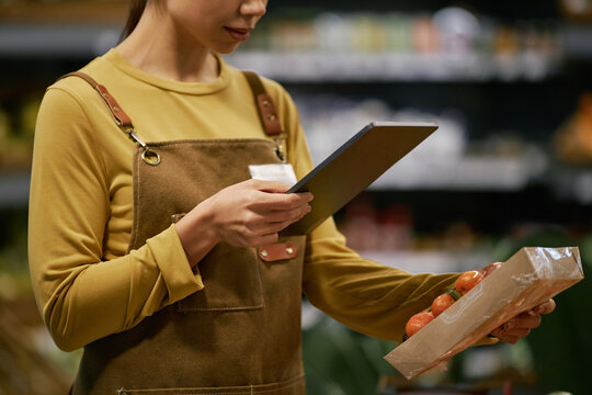 Woman in yellow long-sleeve shirt checking inventory list on digital tablet while holding package with other hand in grocery store environment