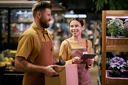 Two grocery store employees smiling and conversing while handling store tasks. Male and female staff members holding a cardboard box and tablet in the bustling supermarket environment