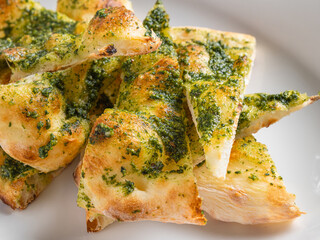 Golden-brown focaccia bread topped with vibrant green pesto, cut into triangular slices and arranged on a white ceramic plate. The crispy crust and airy texture of the bread contrast beautifully with