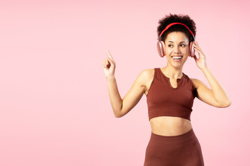 Smiling sporty woman listening to music and pointing finger on pink background