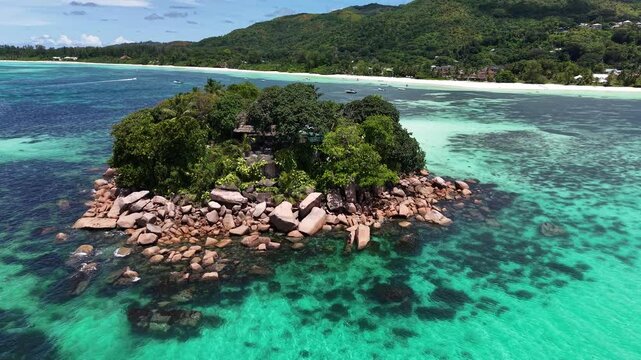 Praslin Island, Seychelles, featuring a tiny rocky islet with palm trees surrounded by crystal-clear turquoise ocean and a nearby sailboat.
