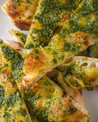 Golden-brown focaccia bread topped with vibrant green pesto, cut into triangular slices and arranged on a white ceramic plate. The crispy crust and airy texture of the bread contrast beautifully with