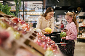 Mother and daughter standing inside grocery store, smiling and selecting fresh fruits together....