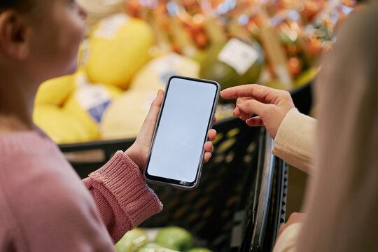 Person showing someone else smartphone in grocery store with several fruits and vegetables visible in background. Engaging in discussion about information displayed on screen