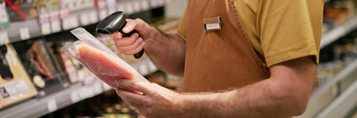 Website header shot of worker scanning package of meat in grocery store aisle, wearing name tag and focusing on task, showcasing efficient inventory management