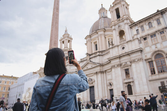 Young woman tourist is taking a photo of sant'agnese in agone and the obelisk using her smartphone in the famous piazza navona in rome, italy