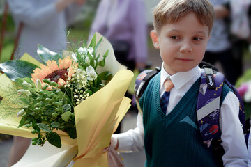A 7-year-old boy carries a bouquet of flowers as he walks to his first day of school on September 1st, dressed in formal attire with a backpack.