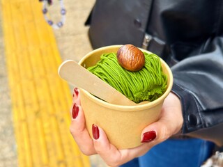 Close-up of a matcha-flavored mont blanc dessert served in a paper cup, topped with a chestnut, held by hand.