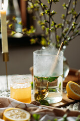 Glass teapot filled with herbal tea containing currant leaves and a golden spoon. A jar of honey and half a lemon are nearby. In the background, a tall candle and a vase with budding branches