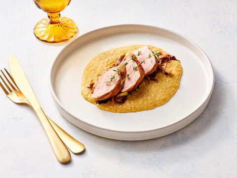 Deliciously plated chicken breast slices lay atop velvety polenta, beautifully garnished with sprigs of fresh herbs. A golden drink complements the dish perfectly