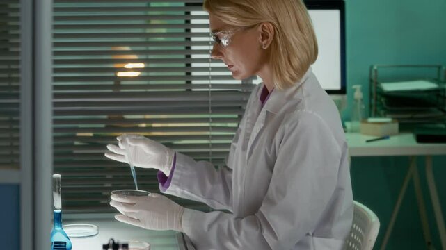 Slowmo shot of concentrated middle-aged Caucasian female chemist in sterile gloves and safety goggles using pipette while mixing liquids in petri dish working in modern research laboratory alone