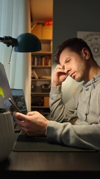 Tired freelancer procrastinating at home office, scrolling phone instead of working. Unproductive man using smartphone at home desk. Concept of procrastination and distraction in remote work