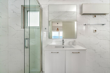 Bright white bathroom with wall-mounted sink, mirror reflecting seaside view, enclosed glass shower, marble tiles, and modern light fixture. Clean, airy, and contemporary space.