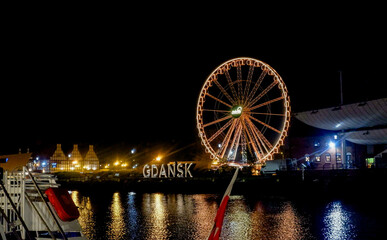 Night view of the illuminated Ferris wheel on Granary Island (Wyspa Spichrzów). The vibrant lights of the Ferris wheel reflect on the Motława River - "GDAŃSK" sign.