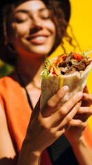 Close-up of doner kebab in young stylish woman's hands with dripping sauce and vegetables against vibrant yellow background, fast food, street food, and delivery