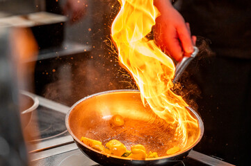 A chef's hand flames up in a frying pan filled with vegetables, creating a dramatic fire effect....