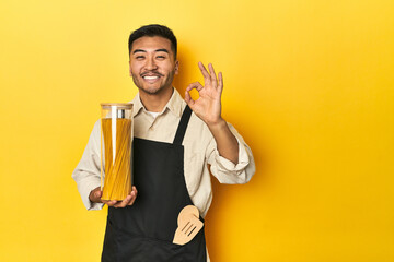 Asian chef with spaghetti jar, yellow studio backdrop cheerful and confident showing ok gesture.