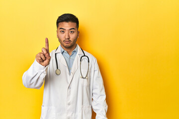 Asian doctor in white coat with stethoscope, yellow studio showing number one with finger.
